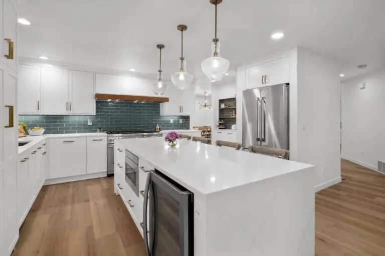 White kitchen with teal subway tile, large quartz island with beverage fridge, brass pendants and stainless fridge