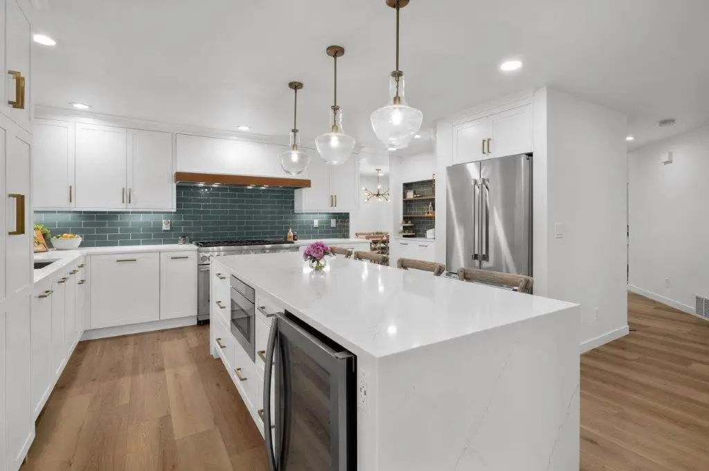 White kitchen with teal subway tile, large quartz island with beverage fridge, brass pendants and stainless fridge