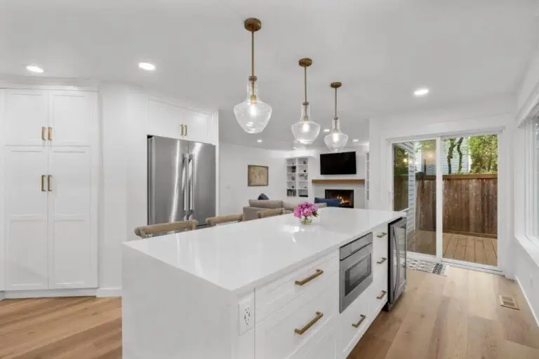 White quartz waterfall island under glass pendants with stainless fridge and view to family room and back deck