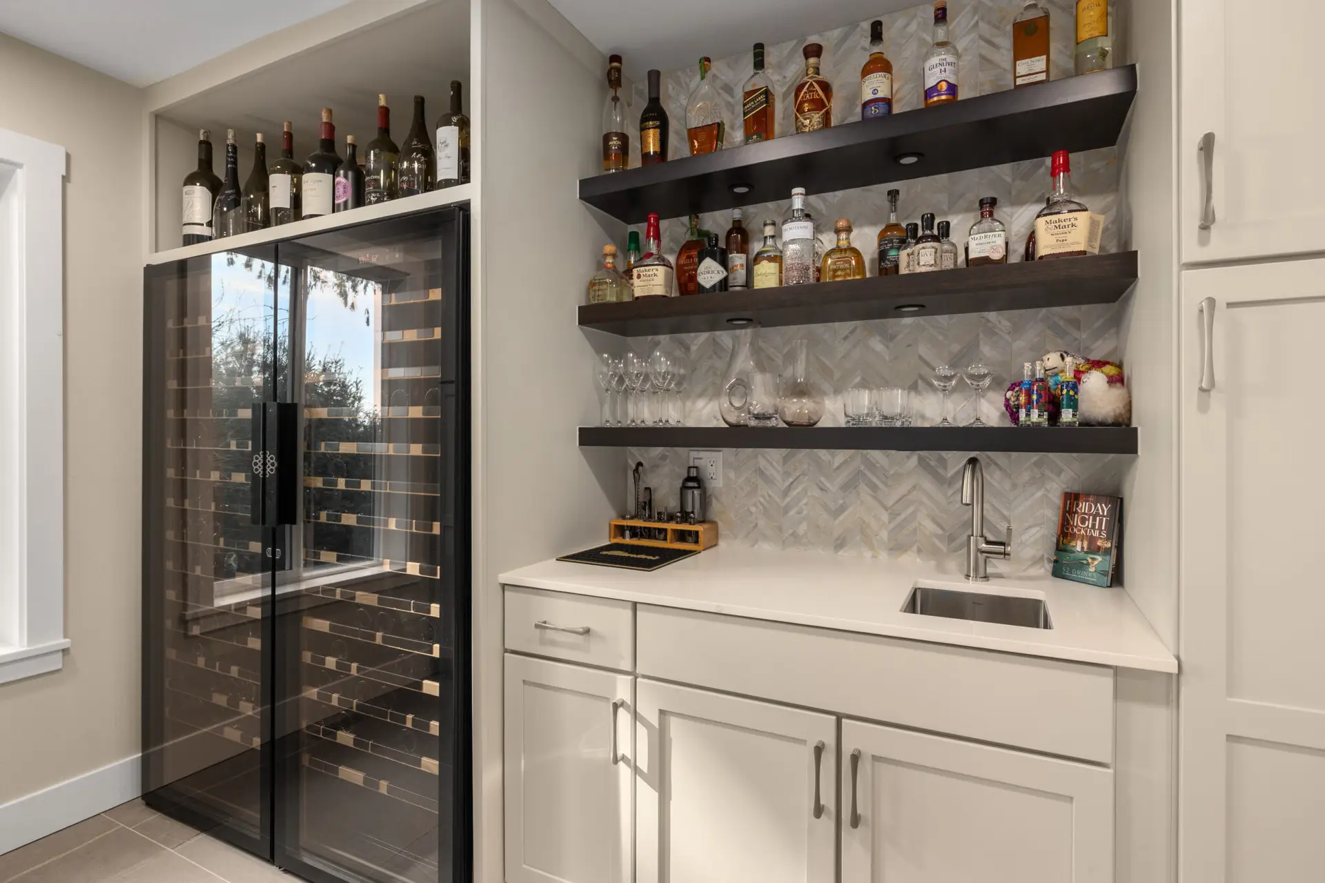 Home bar with glass-door wine fridge, dark floating shelves, herringbone marble backsplash, and undermount sink