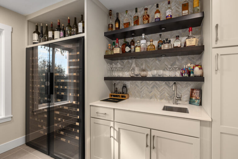 Home bar with glass-door wine fridge, dark floating shelves, herringbone marble backsplash, and undermount sink