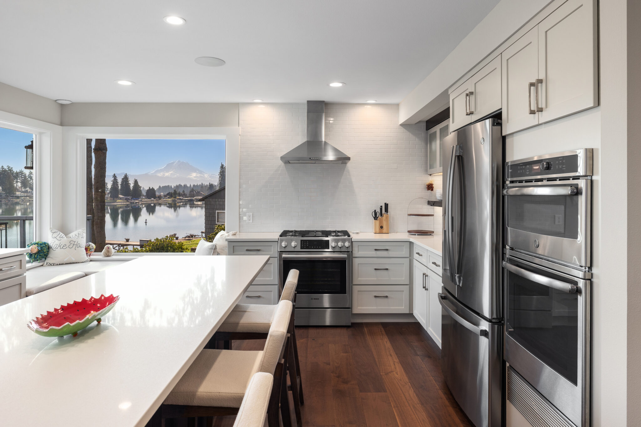 White kitchen with quartz island, stainless double ovens, and a picture window framing Mount Rainier over the lake