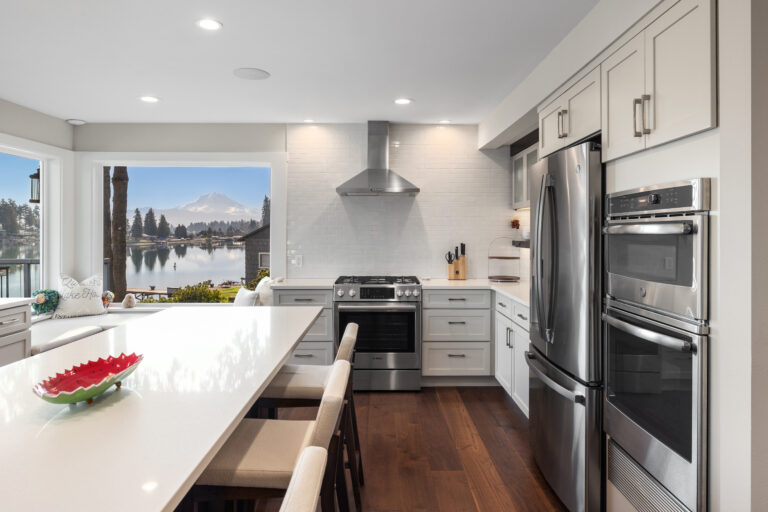 White kitchen with quartz island, stainless double ovens, and a picture window framing Mount Rainier over the lake