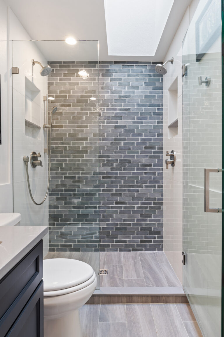 Walk-in shower with blue-gray subway tile feature wall, dual shower heads, tiled niches, and skylight
