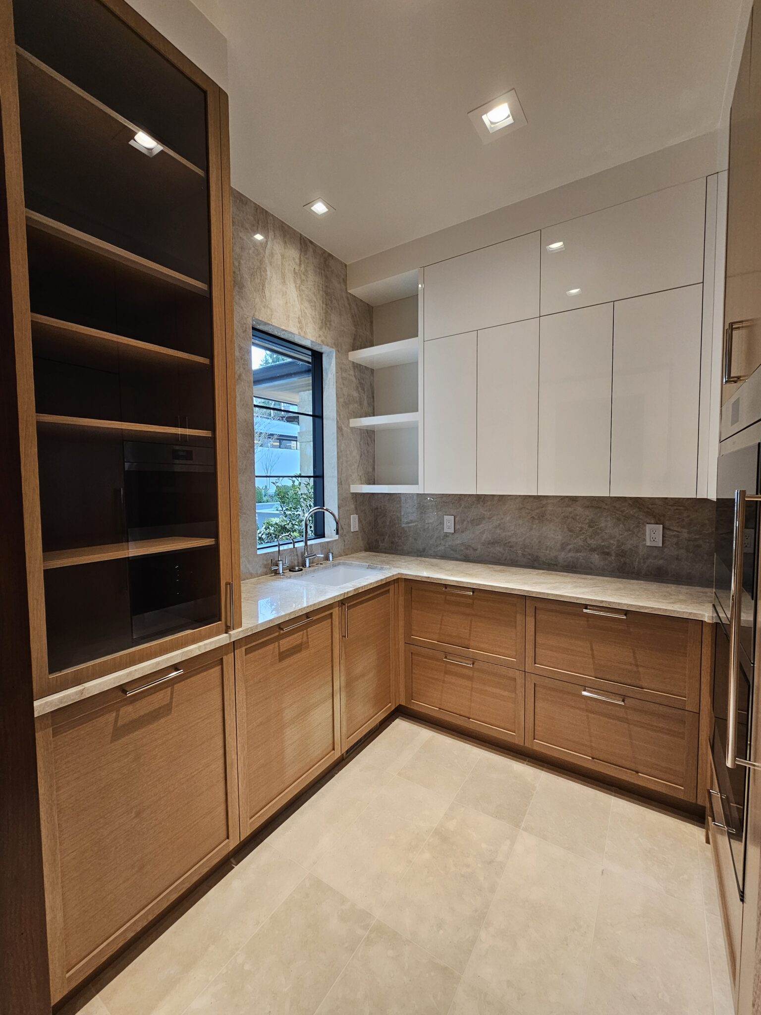 Glass-front oak cabinet and quartzite slab backsplash in a butler pantry with window over the sink