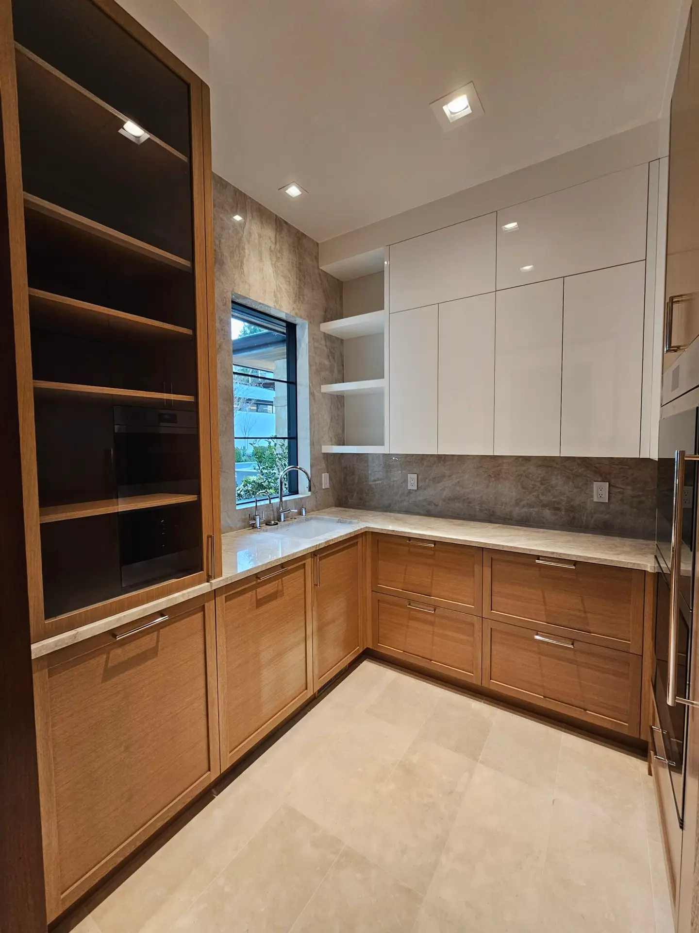 Glass-front oak cabinet and quartzite slab backsplash in a butler pantry with window over the sink
