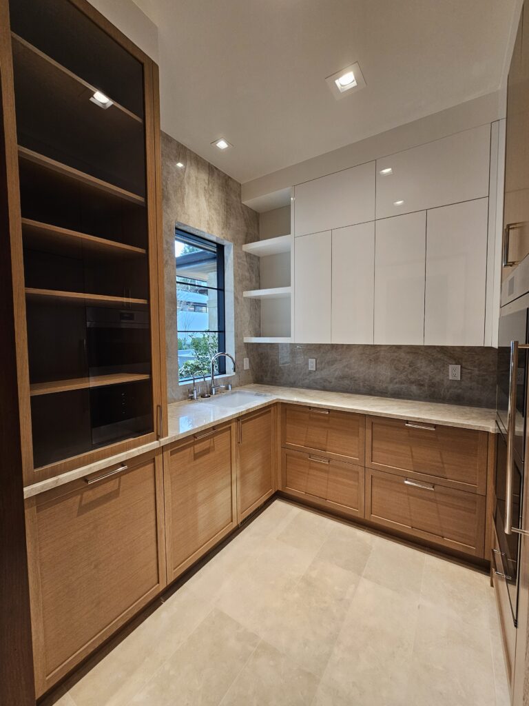 Glass-front oak cabinet and quartzite slab backsplash in a butler pantry with window over the sink