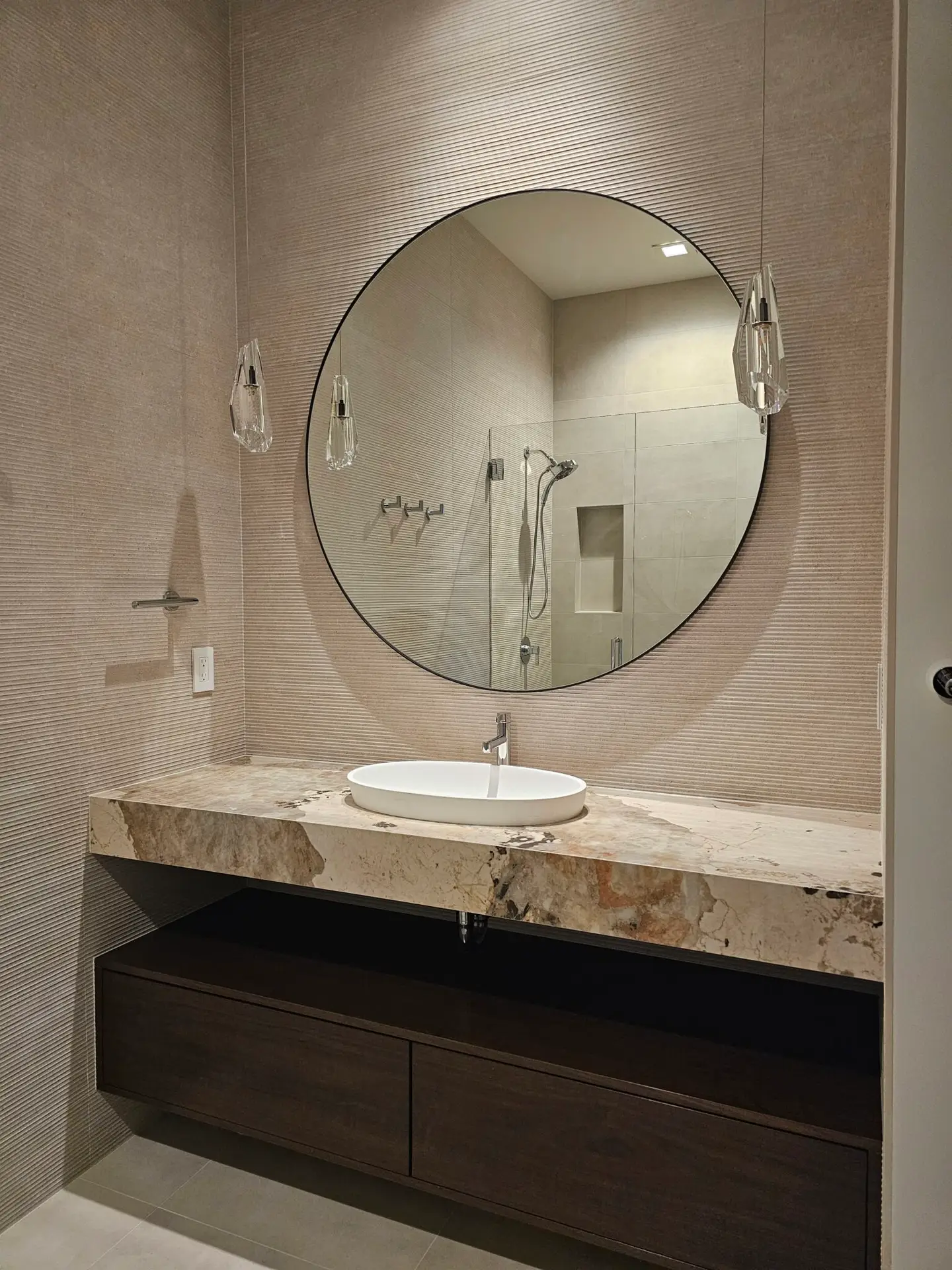 Round mirror and wall-mounted faucet over a vessel sink on a bold-veined stone vanity
