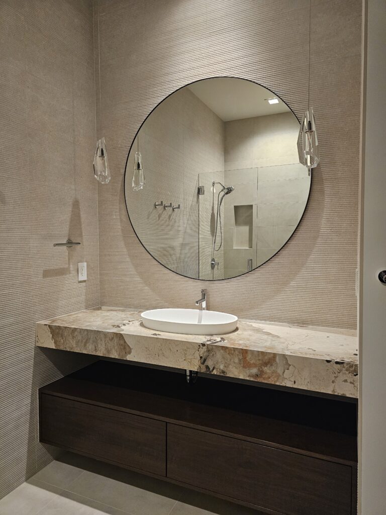 Round mirror and wall-mounted faucet over a vessel sink on a bold-veined stone vanity