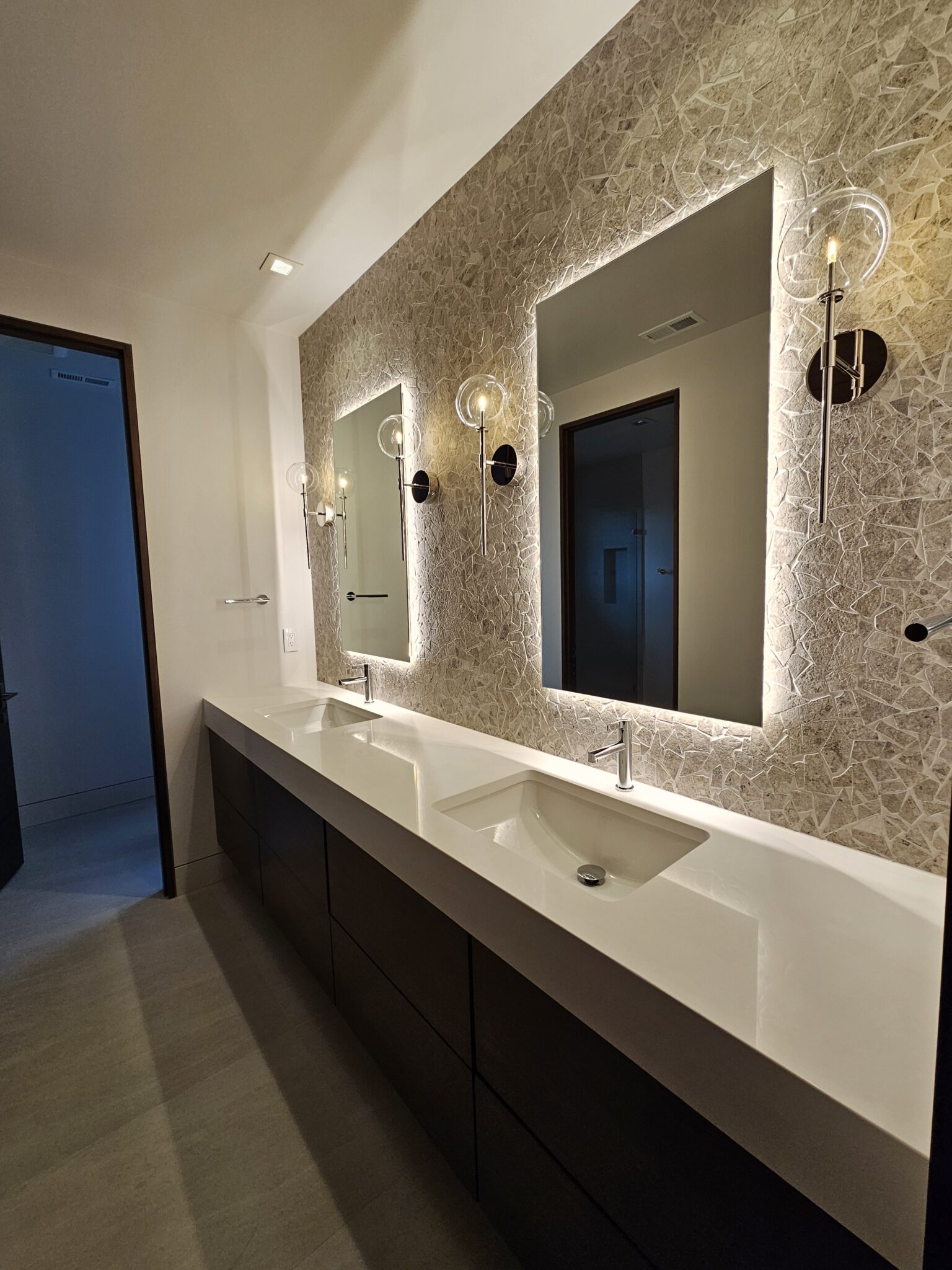 Bathroom vanity with crushed stone mosaic accent wall, backlit mirrors, and wall-mounted faucets