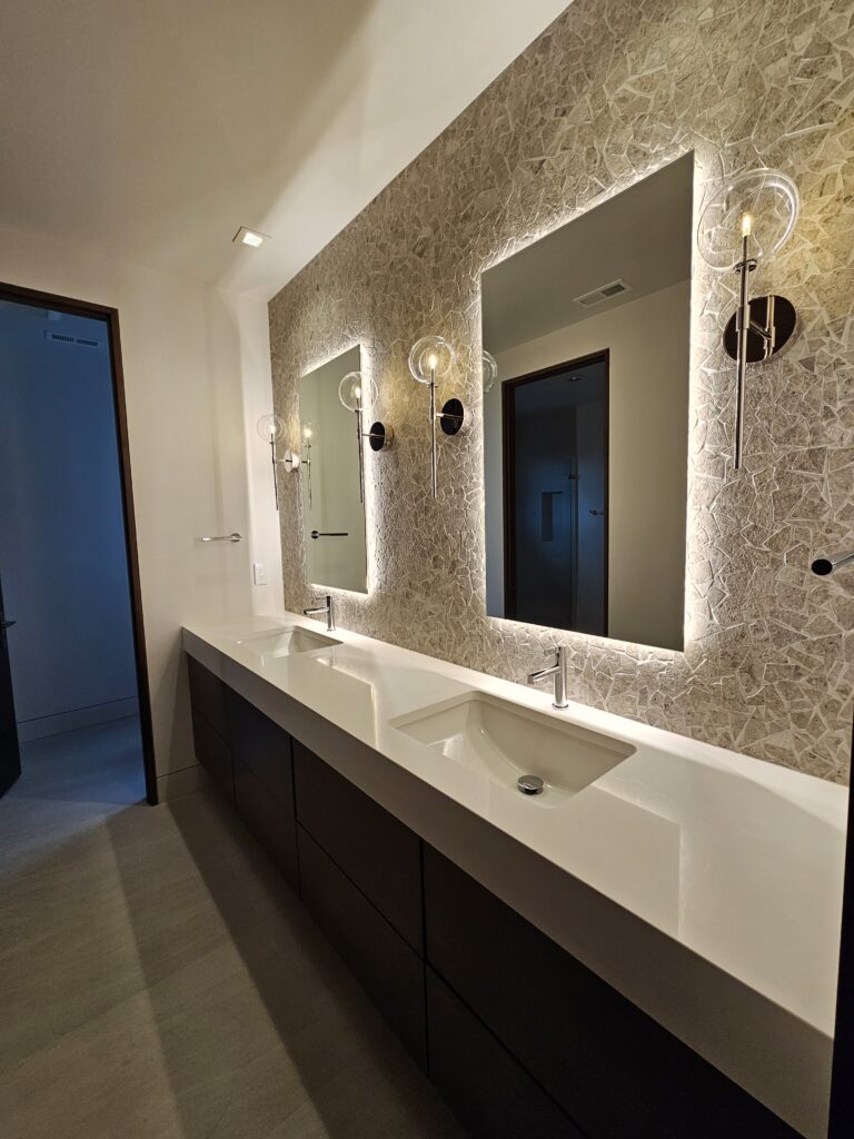 Bathroom vanity with crushed stone mosaic accent wall, backlit mirrors, and wall-mounted faucets