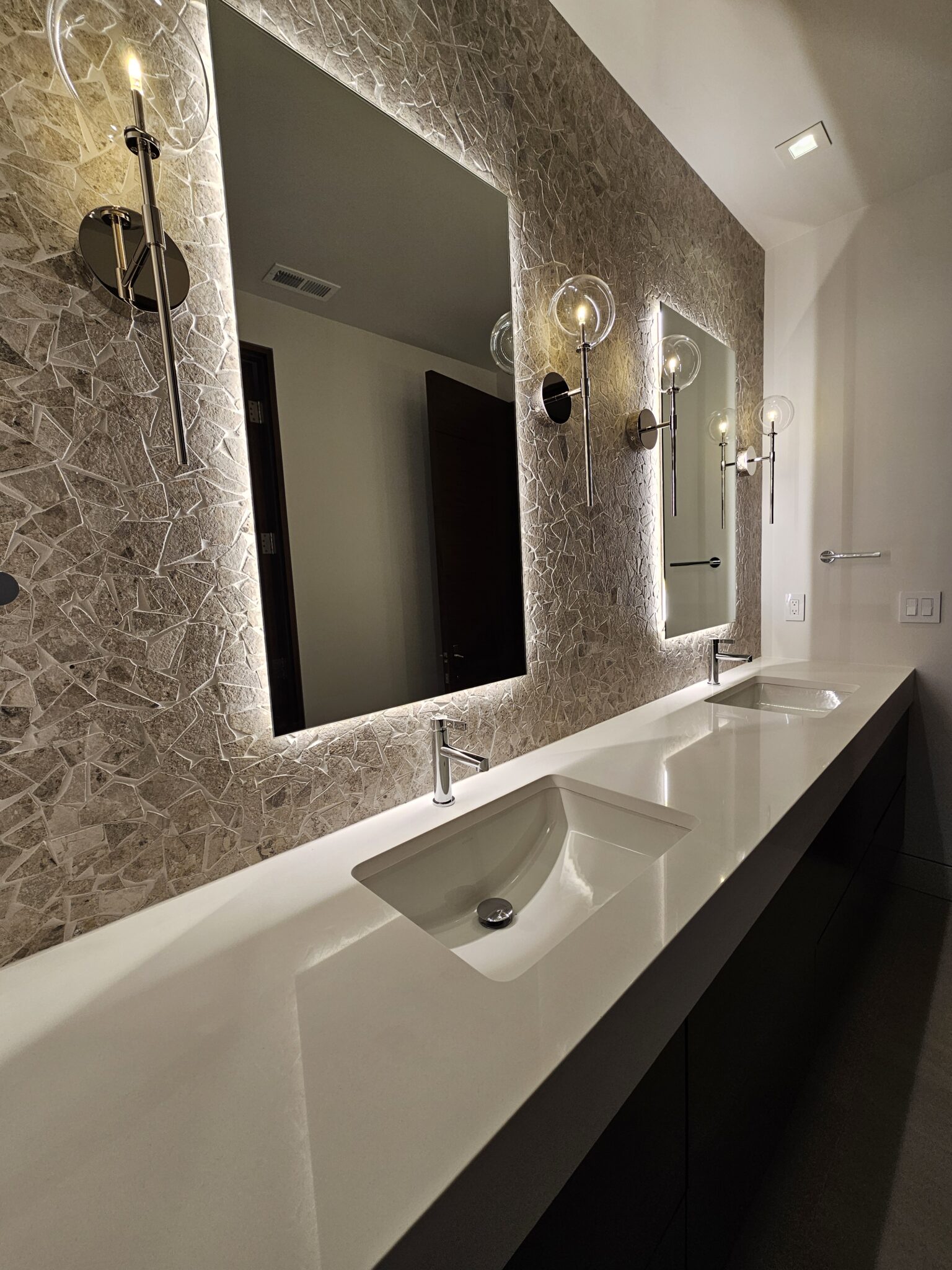 White quartz floating vanity with two backlit mirrors and globe sconces on a stone mosaic wall