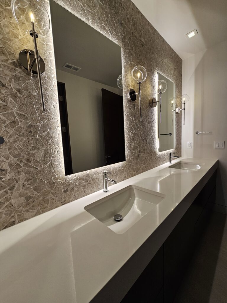 White quartz floating vanity with two backlit mirrors and globe sconces on a stone mosaic wall