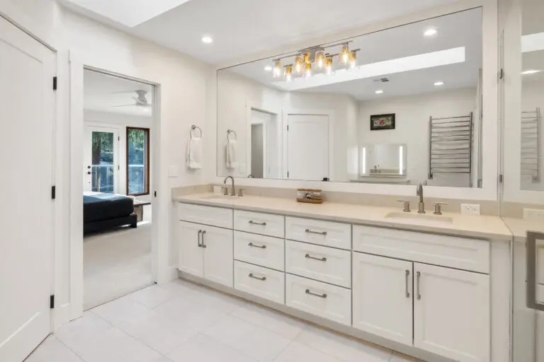 Primary bath double vanity in white with quartz counters, wide mirror, and view into the bedroom