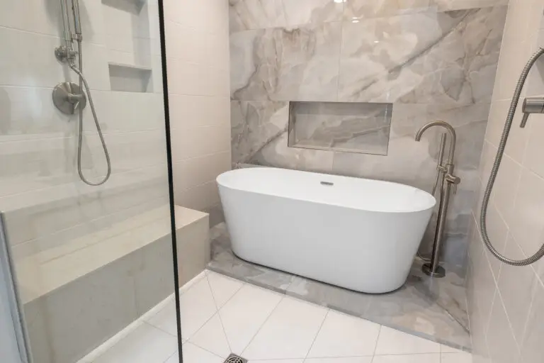 Freestanding white tub beneath a tiled niche in a marble-look wet room with brushed nickel fixtures