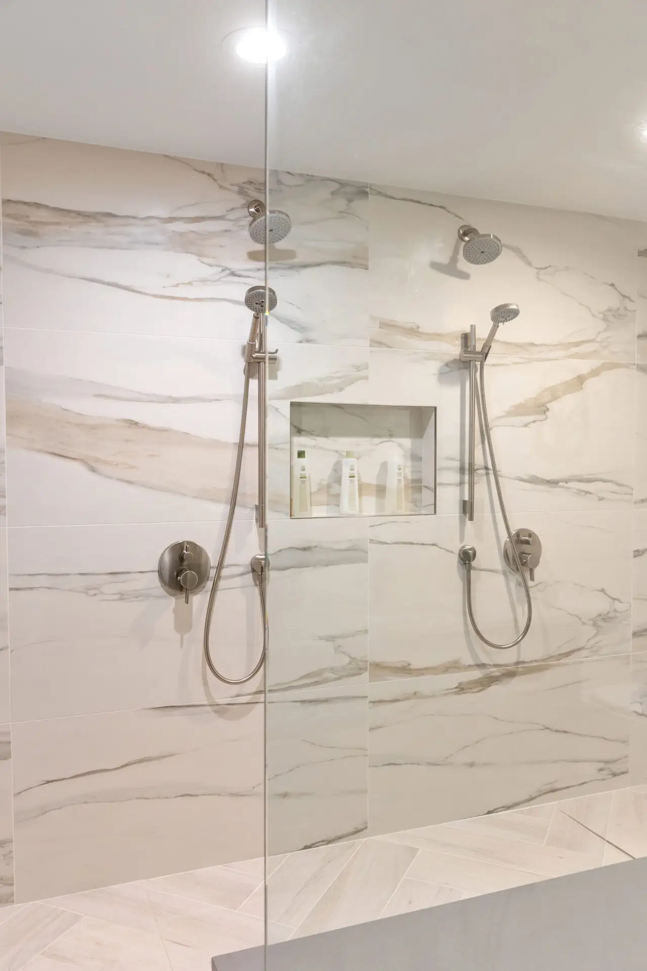 Marble-look tiled shower wall with dual rain heads, hand showers, and recessed niche behind glass