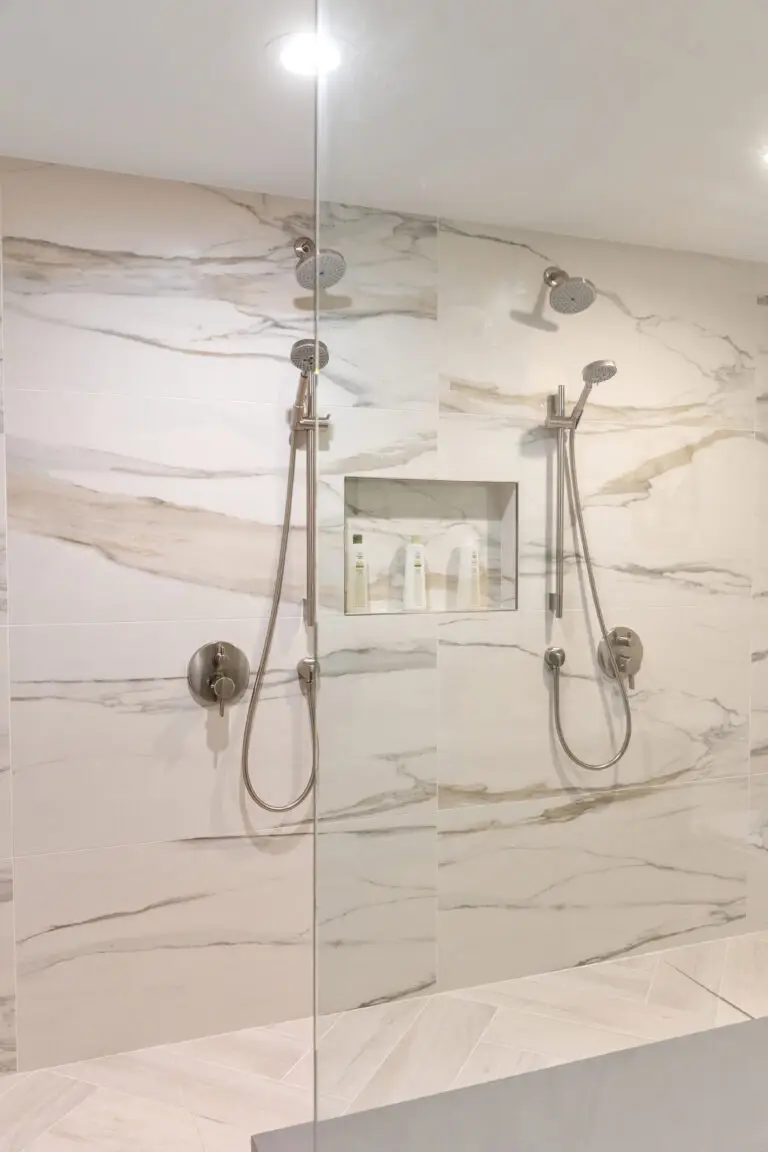 Marble-look tiled shower wall with dual rain heads, hand showers, and recessed niche behind glass
