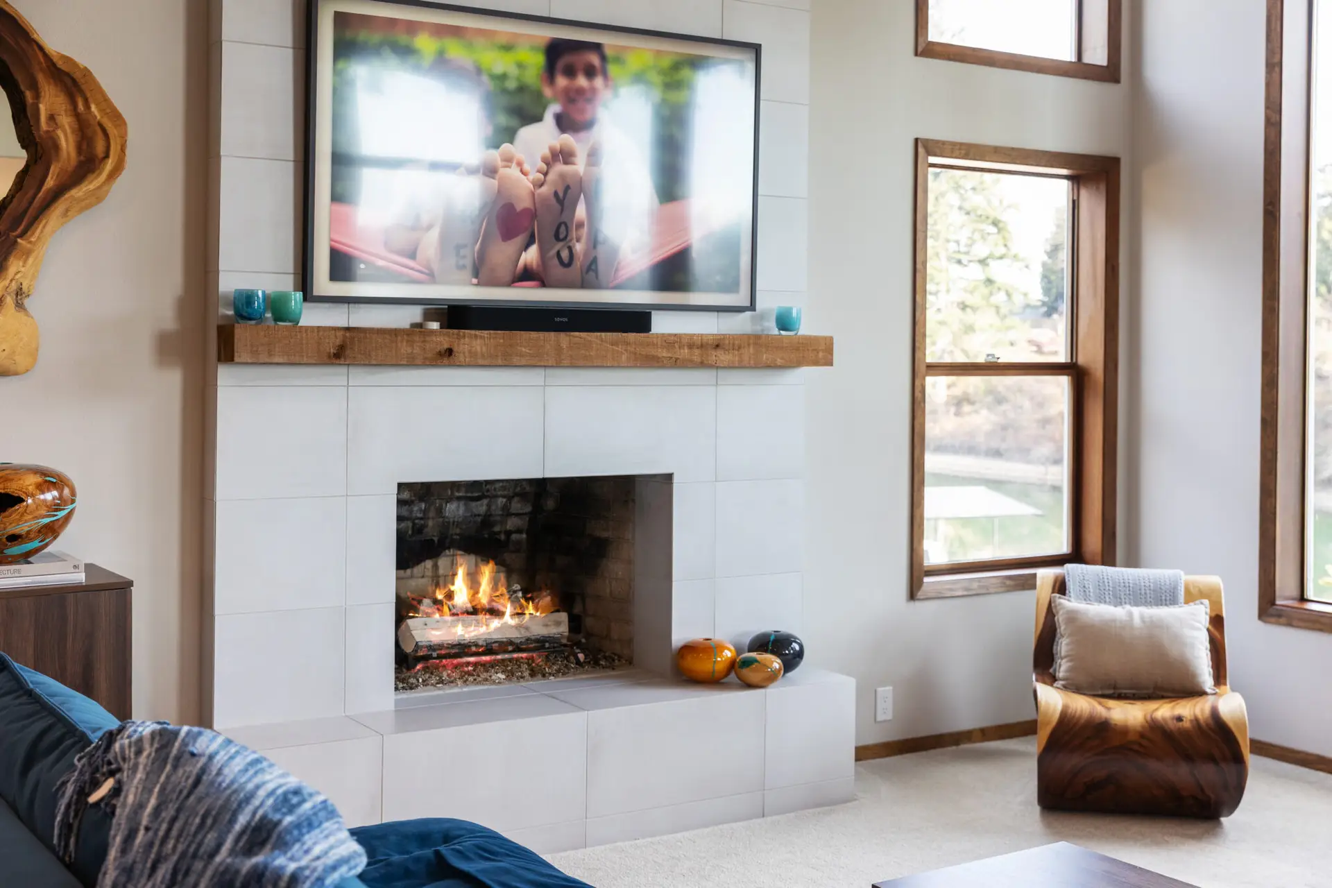 Living room fireplace with white tile surround, rustic wood mantel, TV, and sculptural wood accent chair