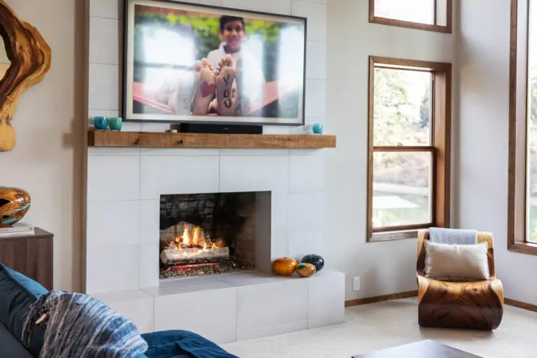 Living room fireplace with white tile surround, rustic wood mantel, TV, and sculptural wood accent chair