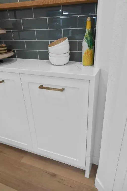 White shaker cabinet with brass pull below a quartz countertop and gray-green subway tile backsplash