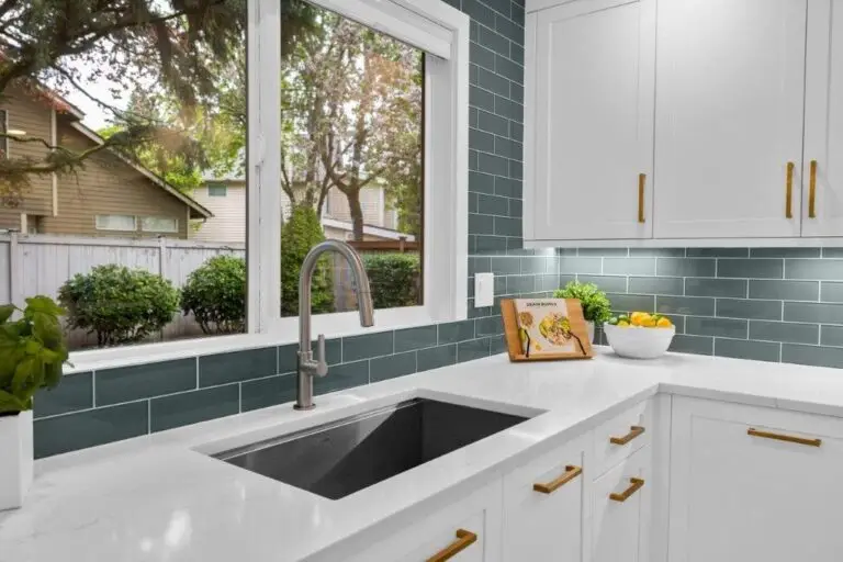 Kitchen sink corner with white quartz counters, teal subway tile to the window sill and brass cabinet hardware