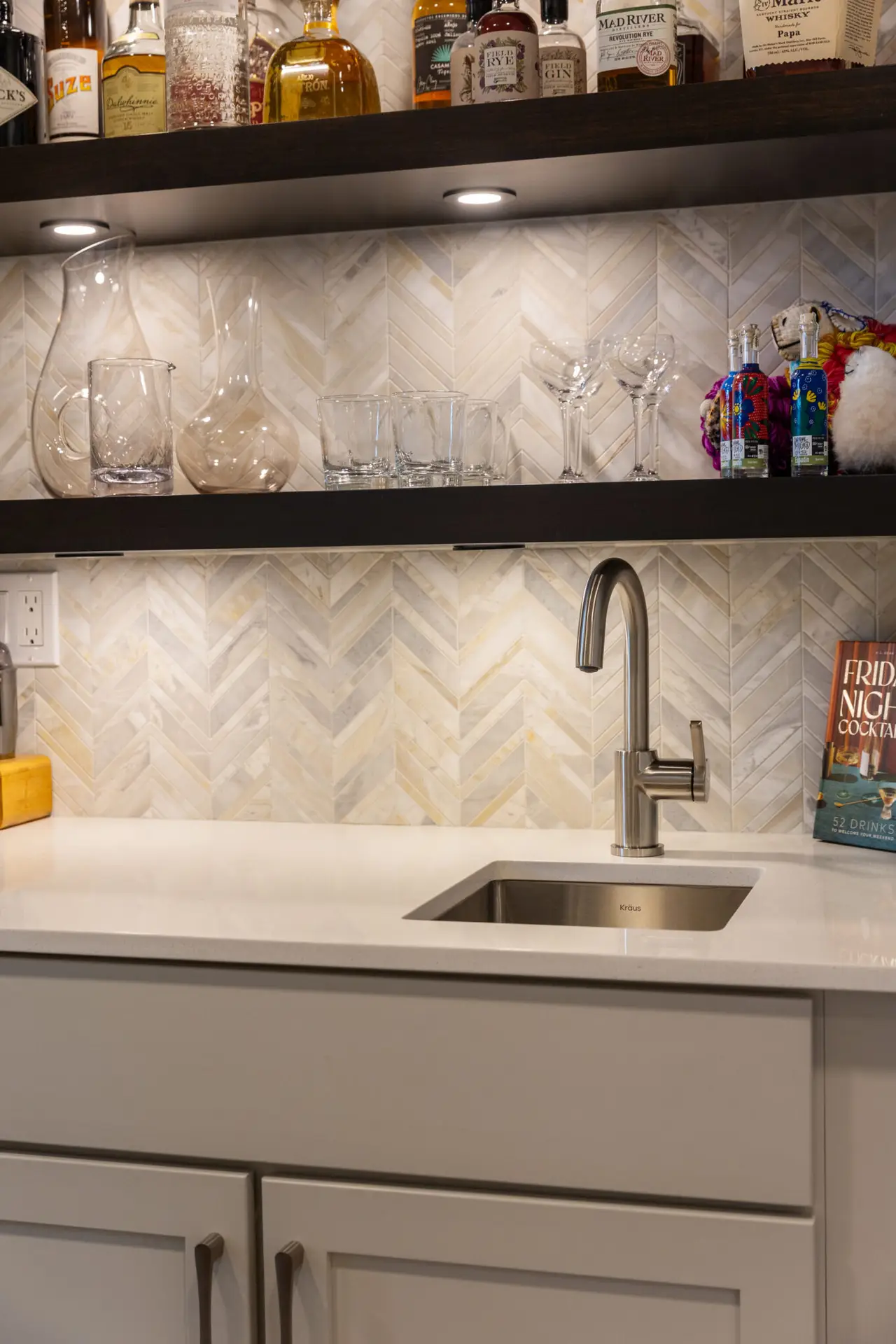 Wet bar detail with chevron marble backsplash, dark lit shelves of glassware, and stainless bar sink