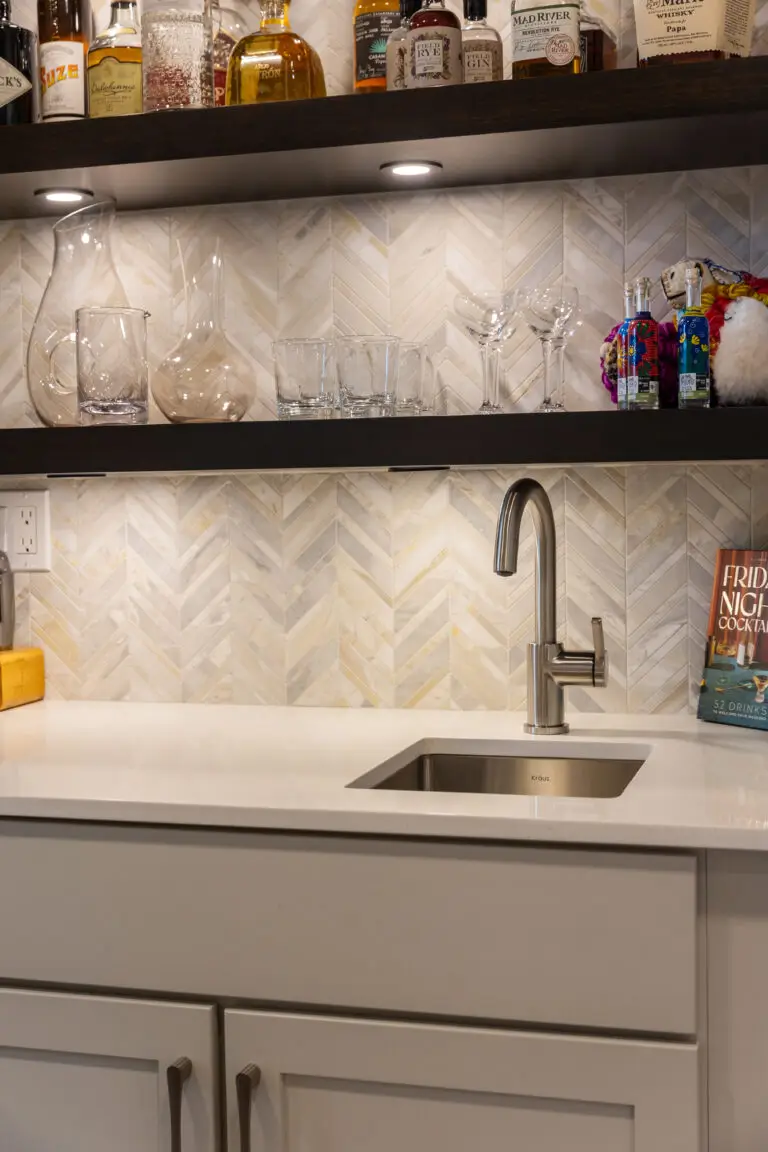Wet bar detail with chevron marble backsplash, dark lit shelves of glassware, and stainless bar sink