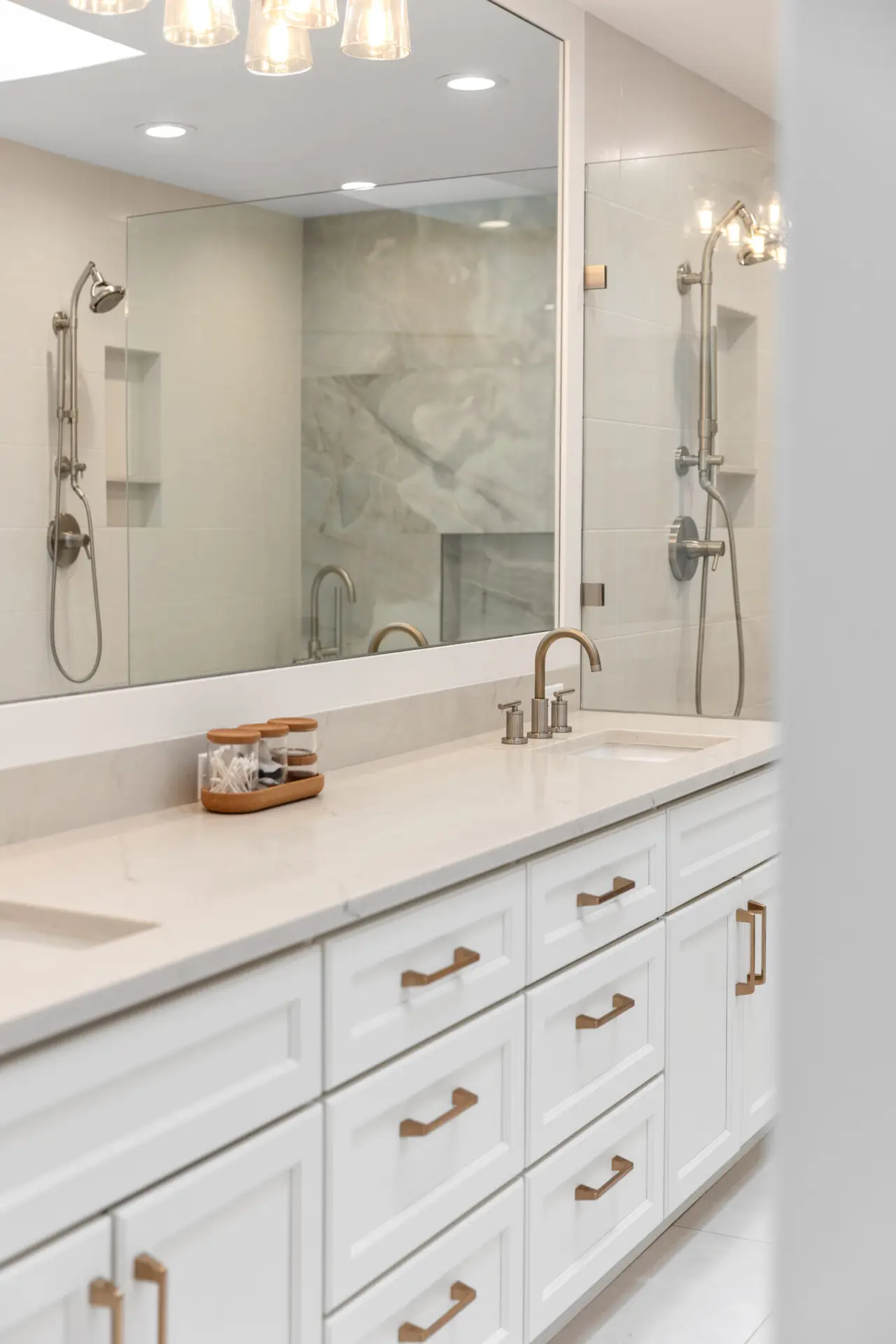 White double vanity with marble-look quartz top, brushed gold pulls, and showers reflected in the wide mirror