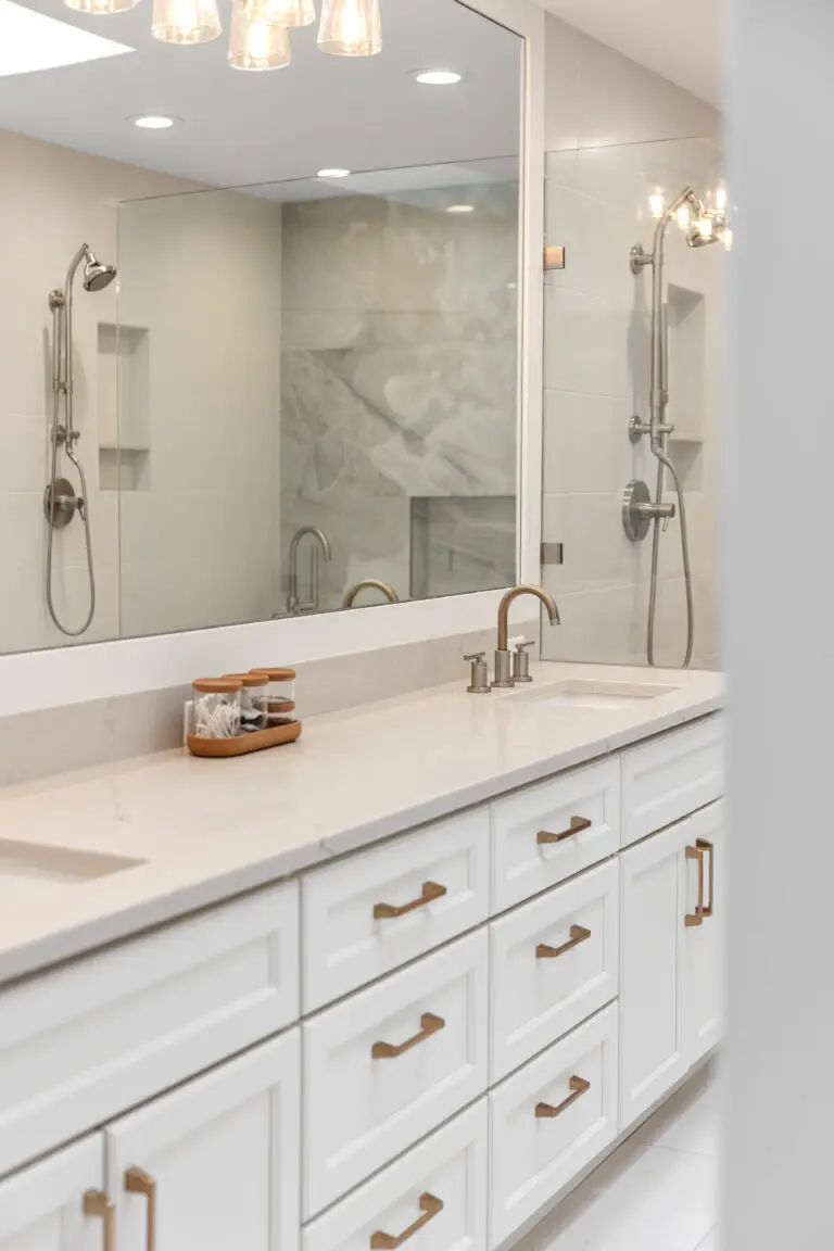White double vanity with marble-look quartz top, brushed gold pulls, and showers reflected in the wide mirror