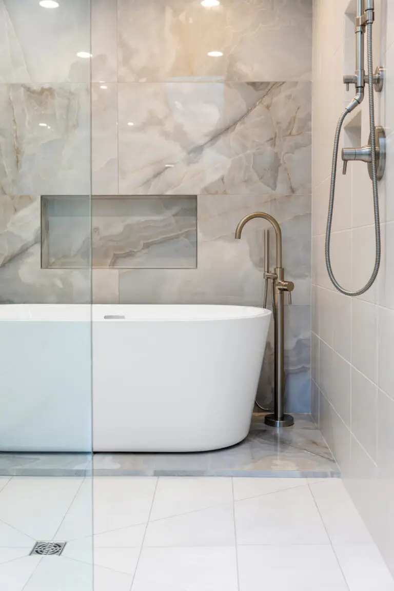 Freestanding white soaking tub with floor-mount brushed nickel filler against marble-look tile wall with niche