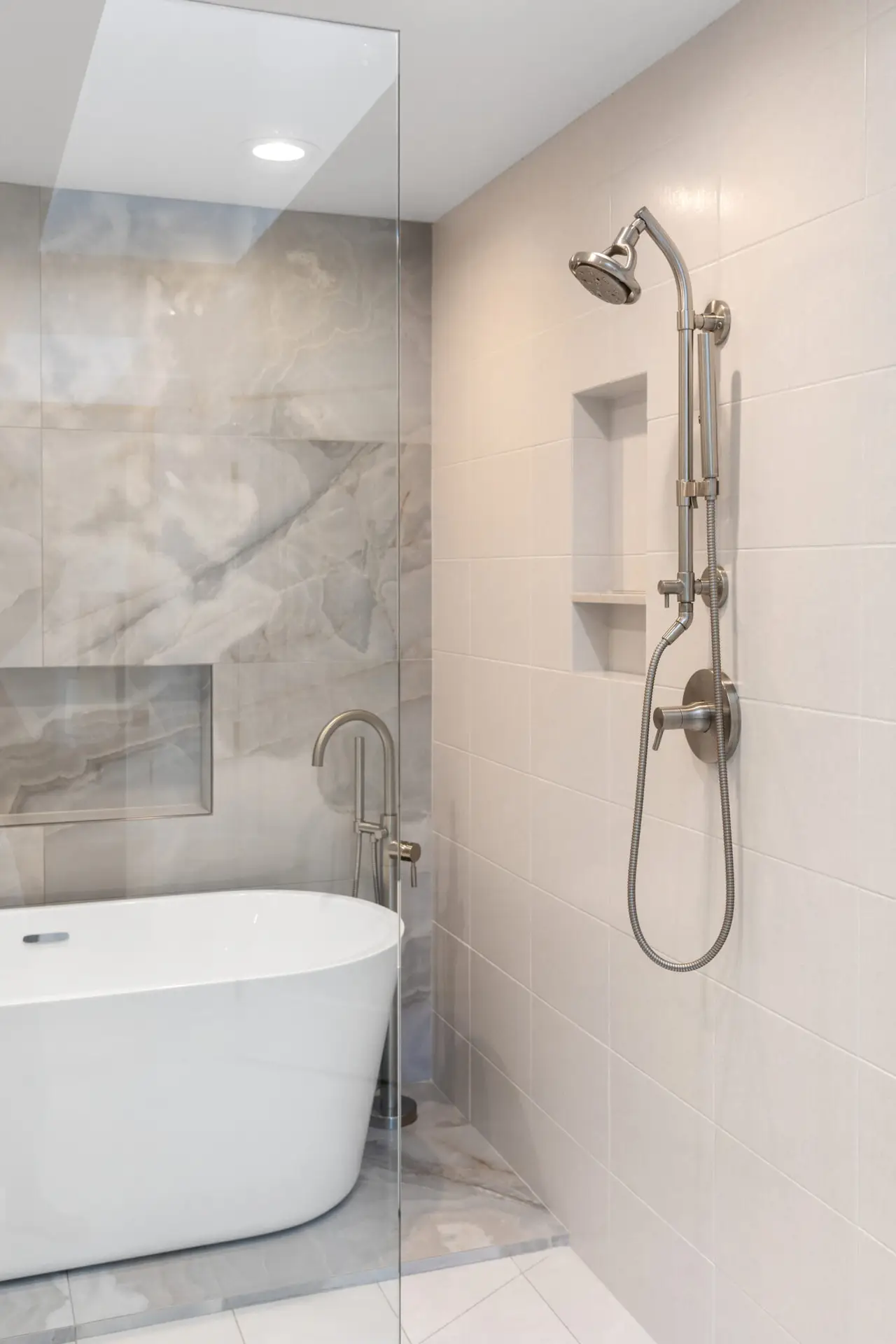 Freestanding tub and marble-look tile wall viewed past a frameless glass panel with wall-mounted shower set