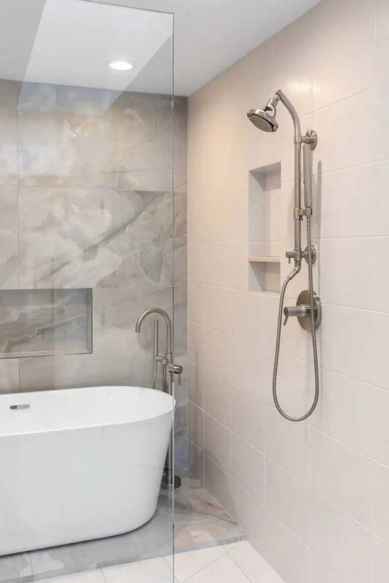 Freestanding tub and marble-look tile wall viewed past a frameless glass panel with wall-mounted shower set