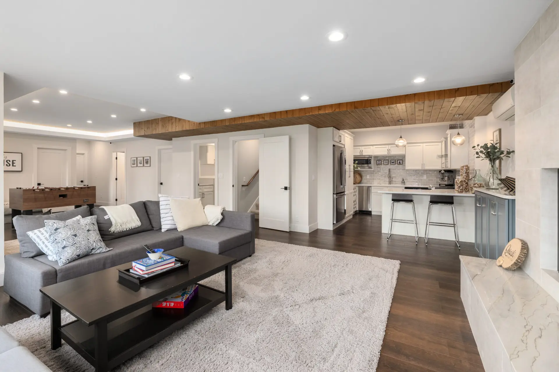 Lower-level great room with gray sectional, foosball table, white kitchenette, and wood ceiling detail