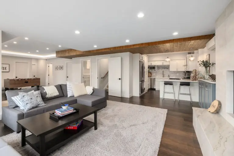Lower-level great room with gray sectional, foosball table, white kitchenette, and wood ceiling detail