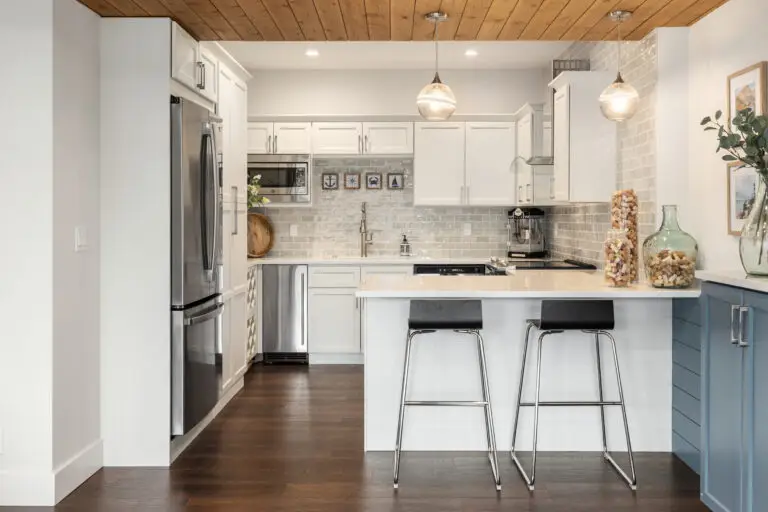 White kitchenette with quartz peninsula, black bar stools, glass pendants, and wood-plank ceiling