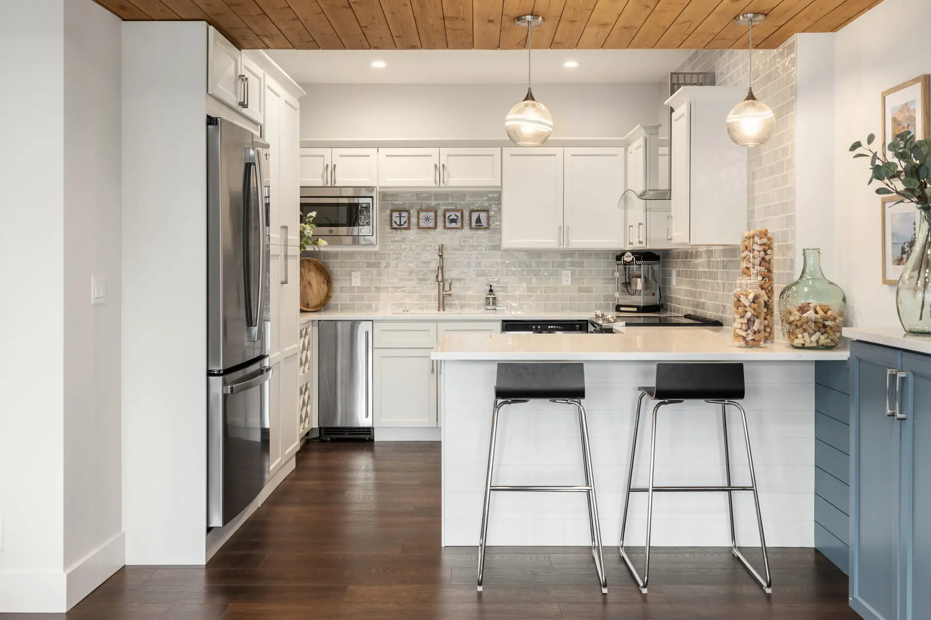 White kitchenette with quartz peninsula, black bar stools, glass pendants, and wood-plank ceiling