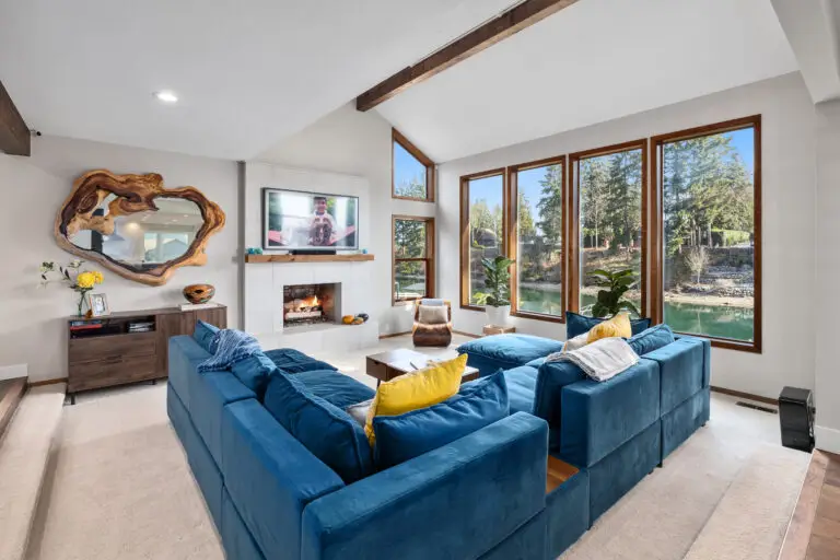 Vaulted living room with blue velvet sectional, tiled fireplace, live-edge mirror, and wood-framed lake windows