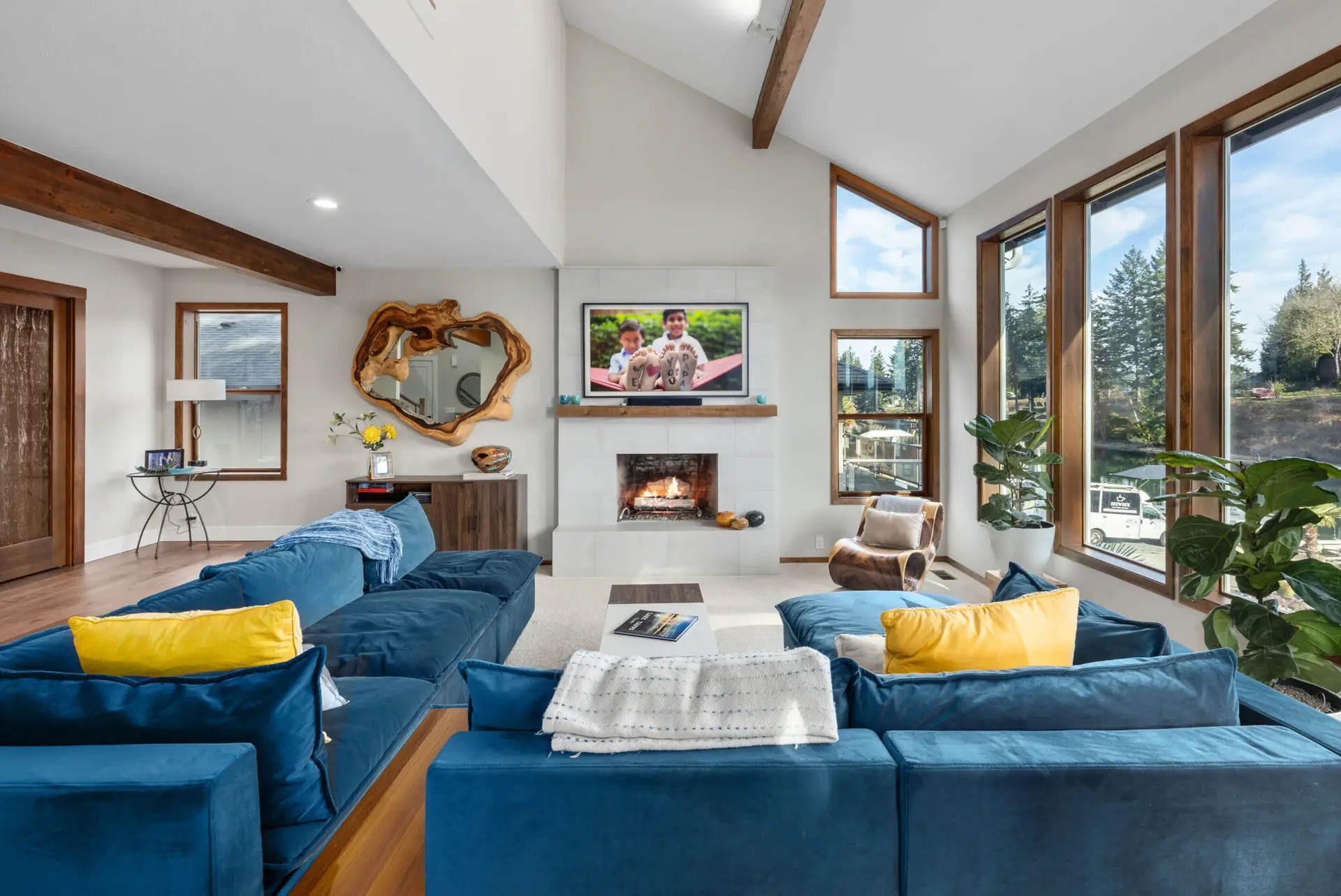 Living room with blue sectional, yellow accent pillows, TV above the fireplace, and live-edge wood mirror