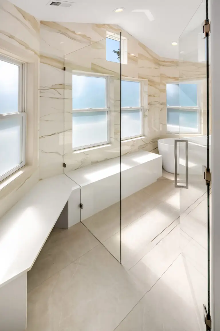 Walk-in shower with glass partition, white bench seating and freestanding tub beyond