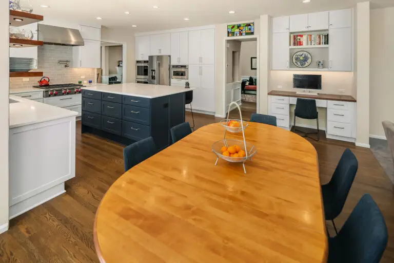 Maple dining table with fruit basket, navy island with white quartz top, and built-in desk in open kitchen