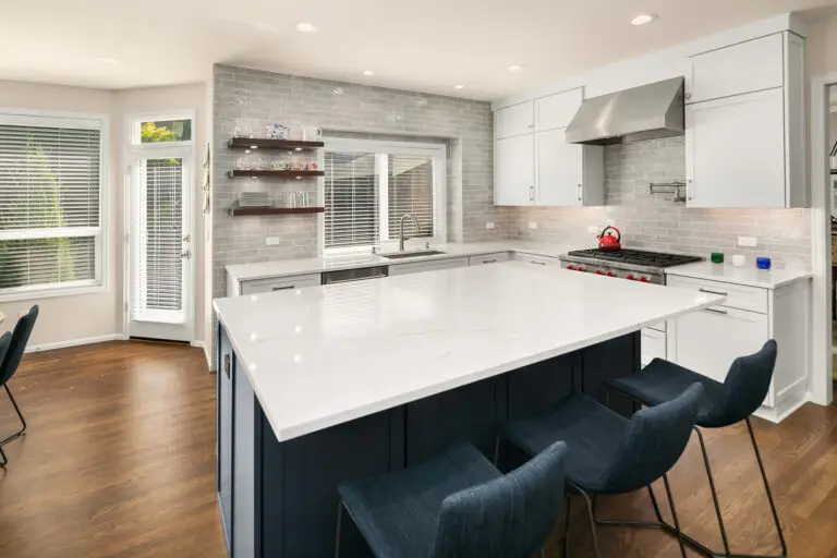 Marble-look quartz island with navy base and denim bar stools in a white kitchen with glossy gray tile