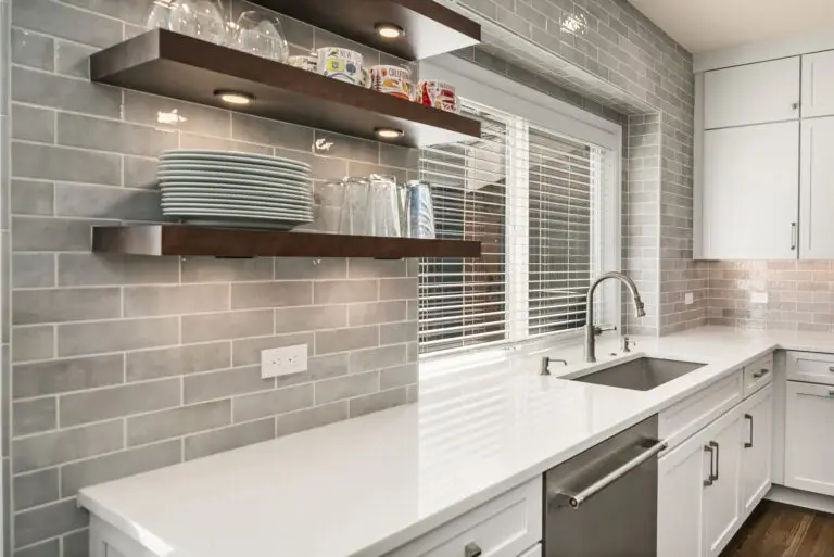 Undermount sink and white quartz counter beneath a window, with lit floating shelves on glossy gray tile