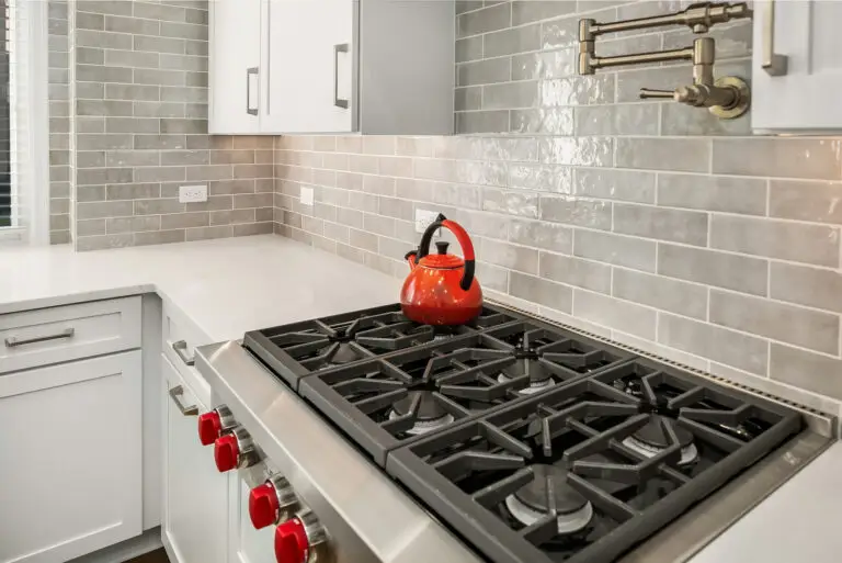 Gas rangetop corner with red kettle, marble-look quartz counters, and shiny gray subway tile backsplash