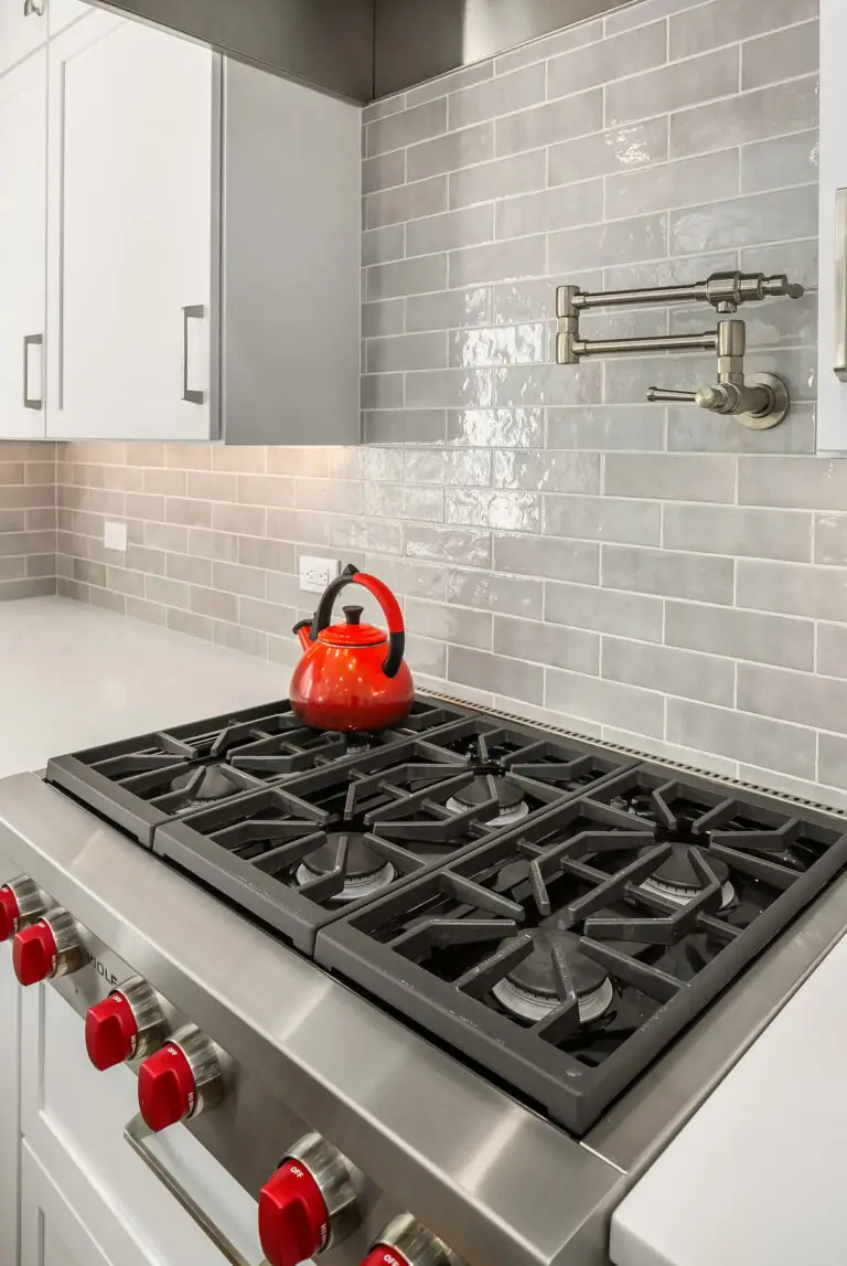 Red kettle on a stainless gas rangetop below a wall-mount pot filler on glossy gray tile