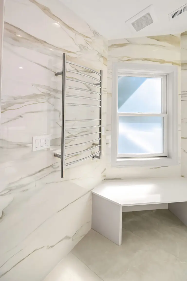 Curved brushed nickel heated towel warmer mounted on marble-look slab wall beside a corner bench