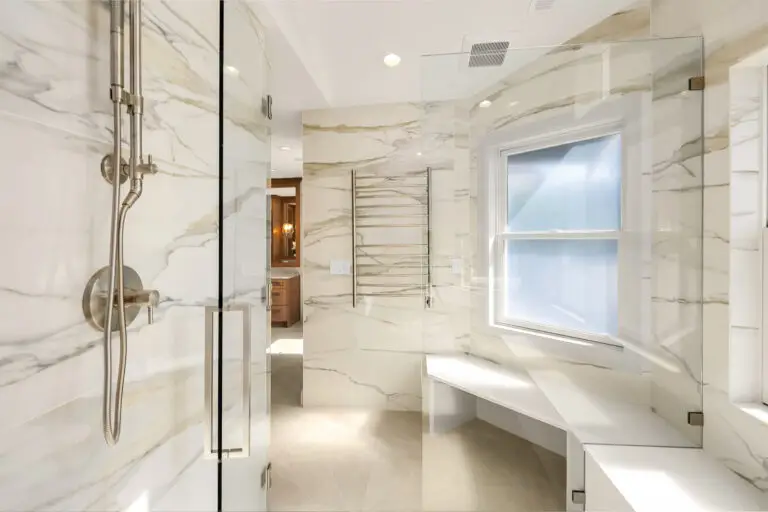Glass-enclosed shower with rain head, corner bench and heated towel rack on marble-look slab walls