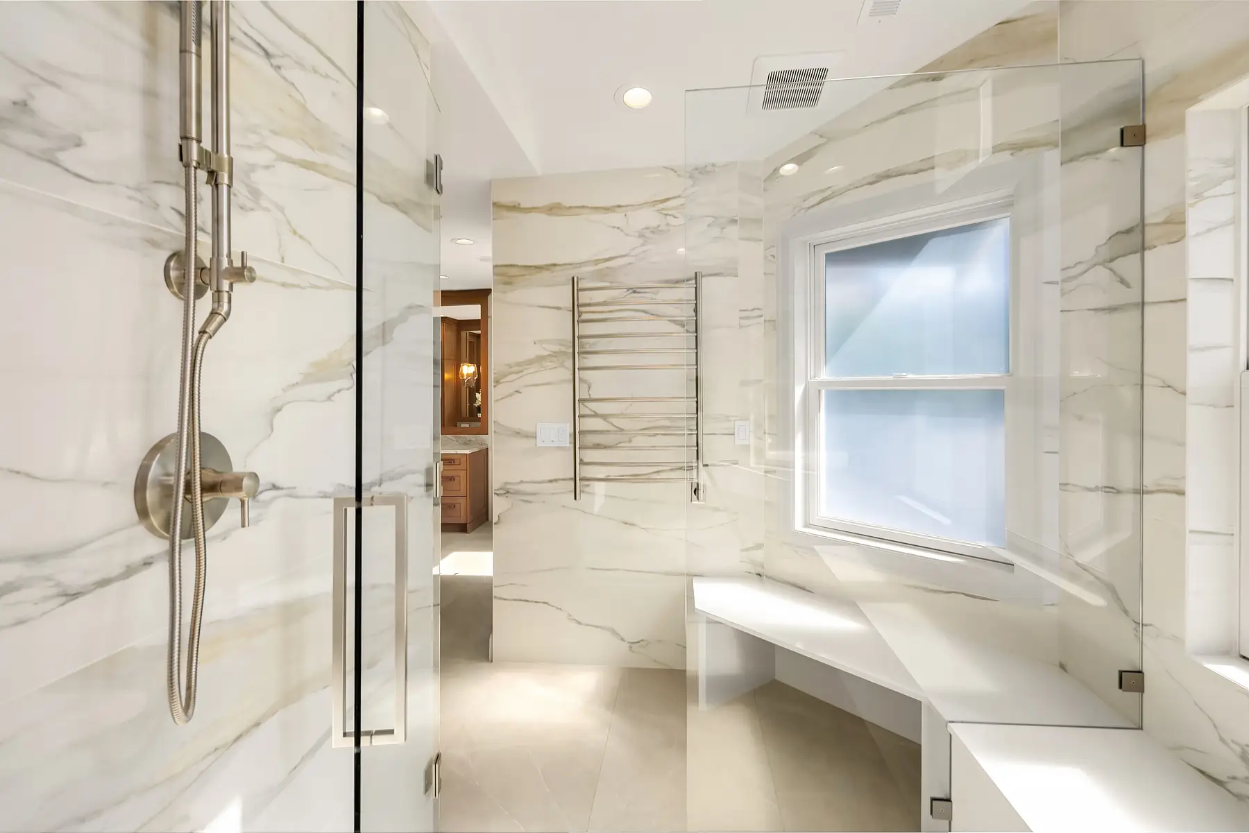 Glass-enclosed shower with rain head, corner bench and heated towel rack on marble-look slab walls