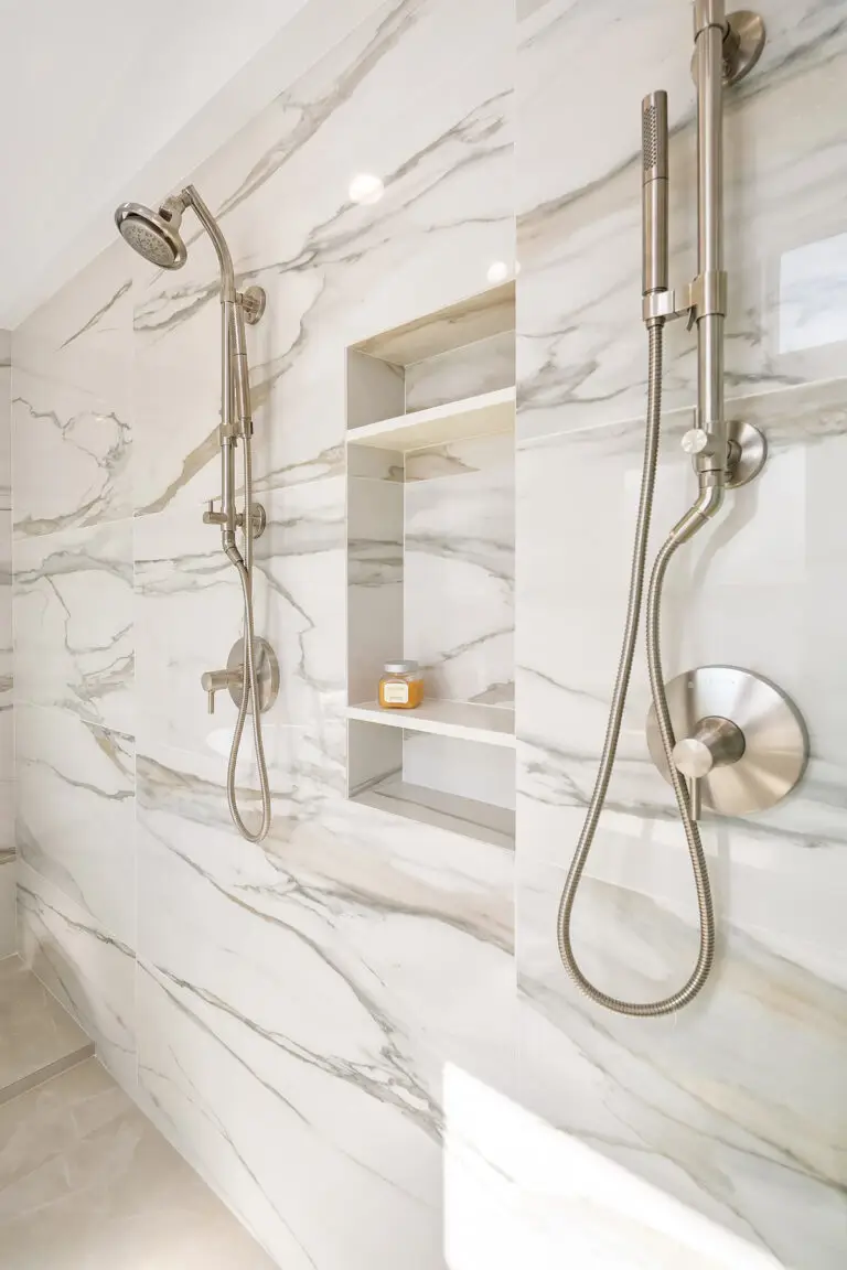 Dual shower heads and handheld sprayer in brushed nickel on marble-look slab walls with tiled niche