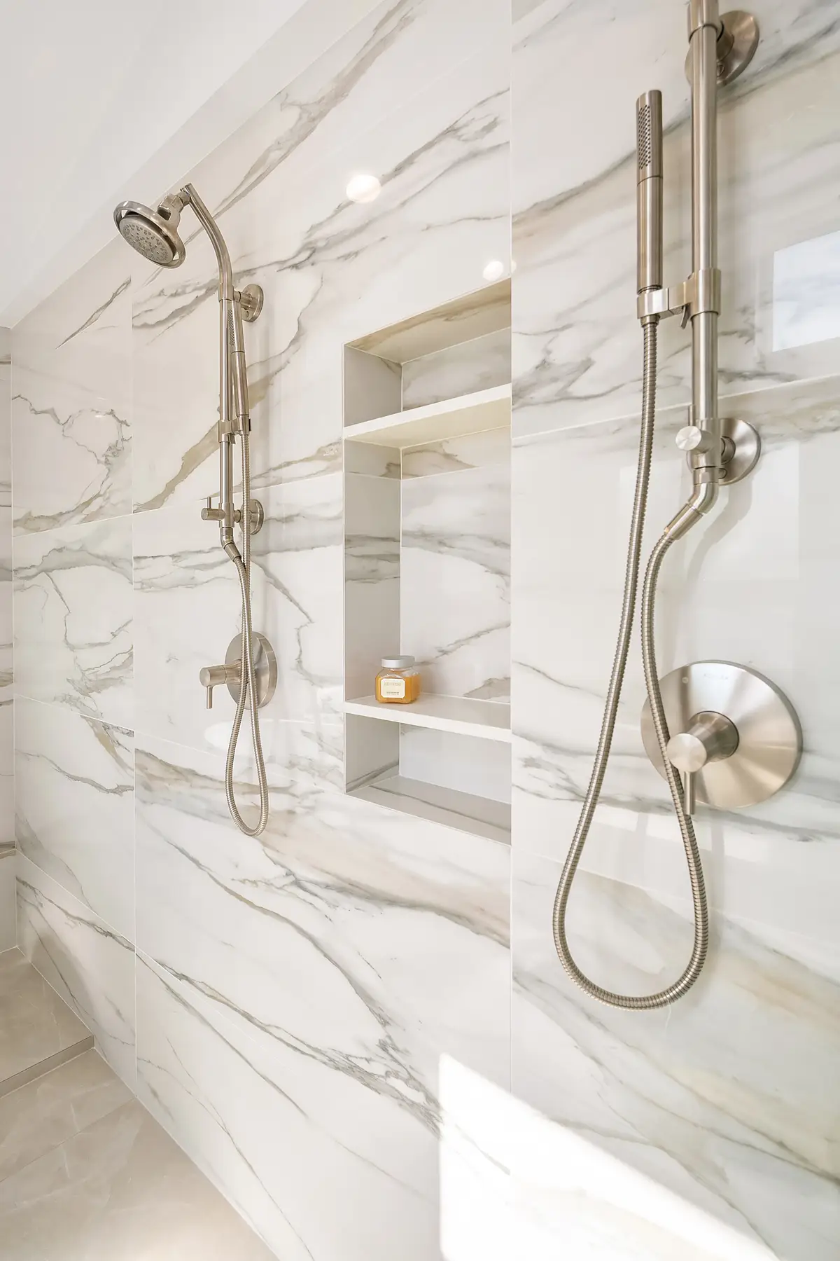 Dual shower heads and handheld sprayer in brushed nickel on marble-look slab walls with tiled niche