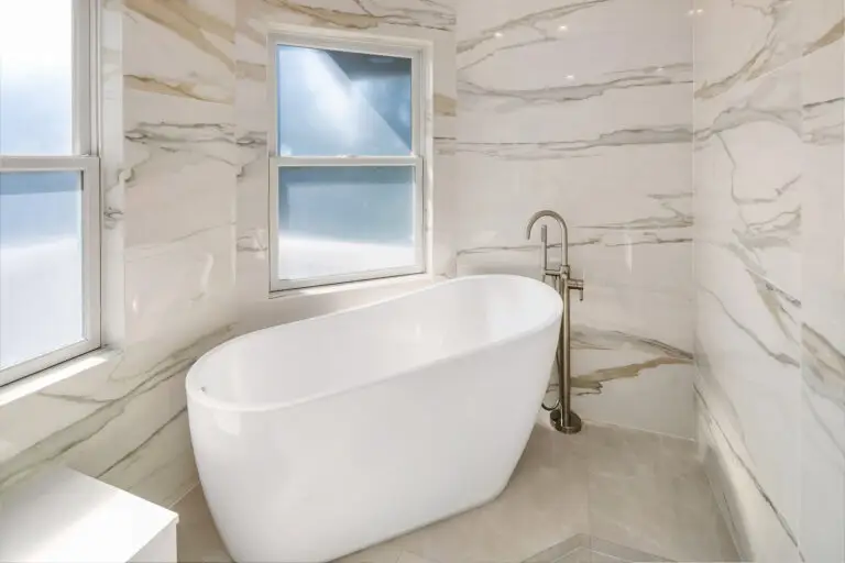 Freestanding white tub beneath a frosted window framed by veined Taj Mahal quartzite-style walls
